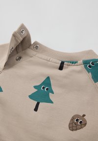 Beige pitkähihaine paita, jossa on vihreitä puita ja ruskeita tammipähkinöitä mustilla silmillä. Sisältää nepparit olkapäässä helppoa pukemista varten.