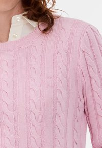 Γυναίκα που φοράει ένα ανοιχτό ροζ πλεκτό πουλόβερ πάνω από ένα λευκό πουκάμισο, με καστανά σγουρά μαλλιά μερικώς ορατά.