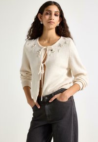 Cardigan in maglia color crema con chiusura a fiocco e decorazioni sulle spalle. Indossato sopra jeans neri a vita alta, con una vestibilità comoda.