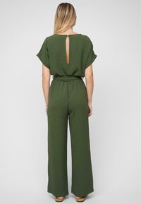 Woman with long blonde hair wearing an olive green short-sleeve jumpsuit with a keyhole back and wide-leg pants, standing against a plain background.
