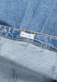 Tessuto di denim azzurro chiaro con una texture leggermente usurata, caratterizzato da un bottone a pressione in metallo e un'etichetta bianca cucita sulla vita.