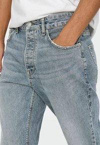 Jeans en denim bleu clair à taille haute, avec poches avant, coupe droite et texture délavée. Détails en métal pour le bouton et les coutures visibles.