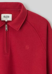 Sweatshirt rouge avec un col zippé, fabriqué dans un matériau doux. Présente un subtil logo "B" en relief sur la poitrine et des manches raglan.
