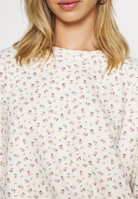 Lehký dlouhý rukáv top v krémové bavlně s květinovým vzorem obsahujícím malé růžové květy a zelené listy; žebrovaný detail výstřihu.