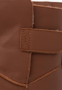 Část hnědého koženého boty s texturovaným povrchem, prošitými detaily a páskem na suchý zip. Na okraji je vidět mírné chlupaté podšívání.