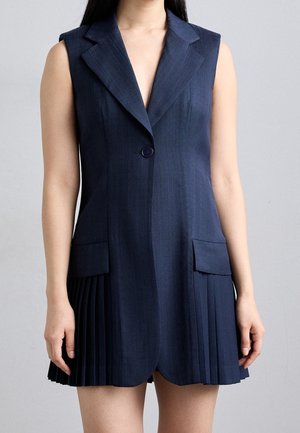 Kobieta w granatowej sukience-żakiecie bez rękawów z rozciętym kołnierzem, pojedynczym guzikiem, kieszeniami z klapkami oraz plisowanymi panelami spódnicy.