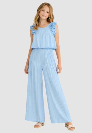 Giovane donna che indossa una tuta senza maniche azzurro chiaro plissettata con spalle arruffate e sandali beige con punta aperta, in piedi davanti a uno sfondo semplice.