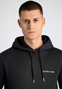 Svart hoodie med ribbstruktur, försedd med en vit broderad logotyp "JUNK de LUXE." Justerbara snoddar med silverdetaljer.