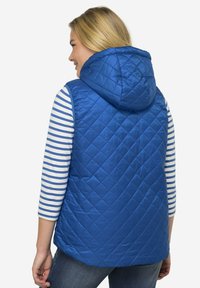 Modrá prošívaná vesta s kapucí, která má hladký, lesklý povrch. Pod ní je pruhovaná košile, která kontrastuje. Neformální design s viditelným šitím.