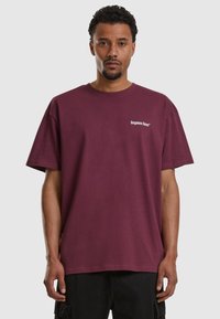 Tricou din bumbac maro, cu mâneci scurte, decolteu rotund și un logo mic alb pe piept, purtat cu pantaloni scurți cargo negri.