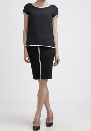 Woman wearing a black short-sleeve top with white trim and a black skirt with a vertical white zipper down the front, paired with black heels.