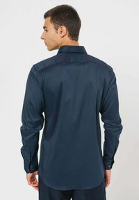 Maglietta a maniche lunghe blu navy in tessuto liscio, con colletto classico, polsini con bottone e orlo arrotondato. Cuciture dettagliate visibili.