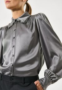 Seidenweiches graues Blusenshirt mit einem Rüschenausschnitt, Knopfleiste und Ballonärmeln. Verfügt über strukturierte Manschetten und eine glatte Stoffreflexion.