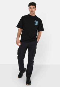 Svart t-skjorte med kort erme som har blå grafisk design på venstre bryst, kombinert med svarte cargo-bukser og svarte sneakers med gummisåle.