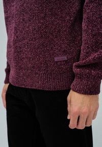 Nahaufnahme einer Person, die einen strukturierten bordeauxfarbenen Pullover mit dem Etikett "Salsa Jeans" am Saum und schwarzen Hosen trägt, mit der Hand entspannt an der Seite.