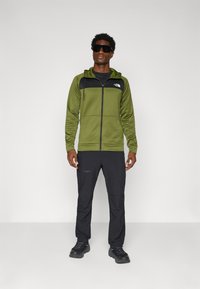 Groene rits hoodie met een zwart paneel, capuchon en voorzakken, gecombineerd met zwarte broek en donkere schoenen. Model draagt een zonnebril.