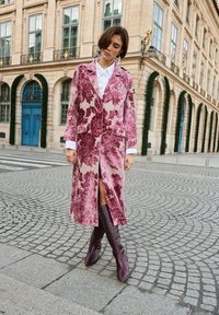 Manteau long à motifs floraux roses et beiges avec des boutons, associé à des bottes marron montantes jusqu'aux genoux et une chemise blanche. Tissu texturé et poches visibles.
