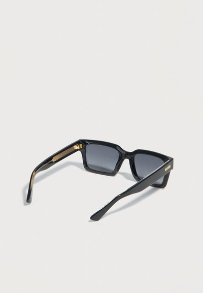 Μαύρα ορθογώνια γυαλιά ηλίου με σκοτεινούς φακούς, χρυσές μεντεσέδες και ένα μικρό χρυσό λογότυπο στο άκρο του βραχίονα, σε απλό φόντο.