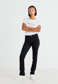 Jeans de mezclilla negra con corte ajustado, que cuentan con un diseño clásico de cinco bolsillos y cierre de botón. Combinados con una camiseta gráfica blanca y zapatillas.