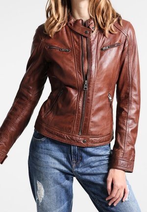 Femme portant une veste en cuir marron ajustée avec des poches zippées et un jean bleu délavé sur un fond blanc uni.