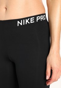 Czarne legginsy Nike Pro charakteryzują się gładkim, elastycznym materiałem oraz szerokim białym paskiem na talii z nadrukowaną nazwą marki.