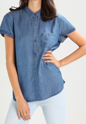Vrouw draagt een korte mouw blauwe denim blouse met knopen en voorzak en lichtblauwe broek, poseert met één hand op de heup.