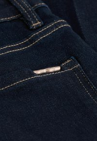 Tessuto denim scuro che mostra una vista ravvicinata di una tasca con cuciture beige e un piccolo accento metallico sul bordo della tasca.