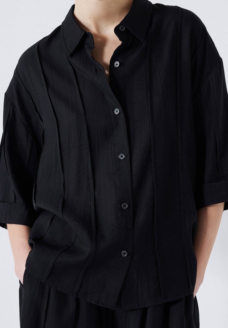 Chemise noire oversize en tissu texturé, à rayures verticales, boutonnée sur le devant et manches retroussées. Dotée de boutons noirs assortis.
