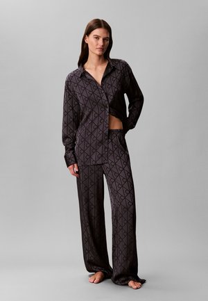 Femme portant une chemise ample noire à motifs et un pantalon large assorti, debout pieds nus devant un fond gris.