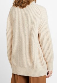 Beige gestrickter Pullover mit vertikaler Rippung, lässiger Passform, langen Ärmeln und weicher Textur. Verfügt über seitliche Taschen und einen leicht abgerundeten Saum.