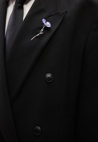 Μαύρο σακάκι διπλής σειράς με ασημένιο λουλουδένιο μπροκέ με ένα λεβαντόχρωμο λουλούδι και αλυσίδες στη γιακά. Συνδέεται μαύρη γραβάτα.