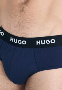 Dunkel marineblaue Unterhosen mit einem schwarzen Bund, auf dem der Markenname "HUGO" in weißem, sich wiederholendem Text zu sehen ist. Glatte Textur und enge Passform.
