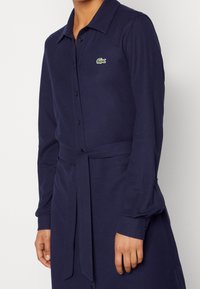 Mörkblå långärmad klänning med krage, knappstängning och ett knytbälte i midjan. Har en liten grön logotyp på bröstet.