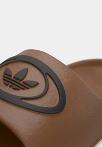 Καφέ σαγιονάρα από καουτσούκ με υφή στην επιφάνεια, με λογότυπο σε μαύρο ανάγλυφο στο λουράκι. Στρογγυλεμένο σχήμα χωρίς ιδιαίτερες διακοσμήσεις.