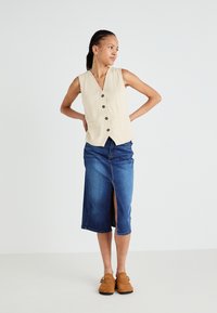 Beige ärmellose Weste mit vorderen Knöpfen, kombiniert mit einem knielangen Jeansrock mit einem Schlitz vorne. Braune Slipper runden das Outfit ab.