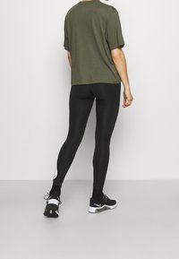 Camiseta de gran tamaño en verde oliva combinada con mallas negras. Las mallas tienen un diseño elegante y un acento blanco cerca del tobillo. Zapatillas deportivas negras.