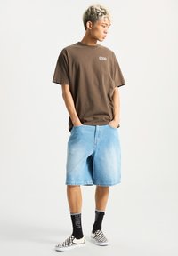 Camiseta de algodón marrón con la palabra "GOOD" impresa en el pecho, combinada con pantalones cortos de mezclilla claros. Calcetines negros con texto blanco y zapatillas de tela de cuadros.