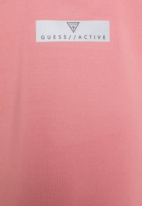 Light pink t-shirt with a smooth texture, featuring a gray rectangular logo that reads "GUESS//ACTIVE" in bold lettering.