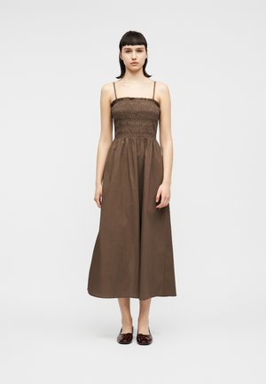 Mujer joven de pie, vestida con un vestido midi marrón sin mangas con cuerpo fruncido, tirantes delgados y zapatos planos de color rojo oscuro, frente a un fondo blanco liso.