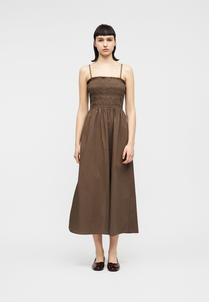 Νεαρή γυναίκα στέκεται φορώντας ένα καφέ μίντι φόρεμα με ριχτό μπούστο χωρίς μανίκια, λεπτές τιράντες και σκούρα κόκκινα μπαλαρίνες, μπροστά σε ένα απλό λευκό φόντο.