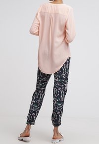 Blusa rosa chiaro a maniche lunghe con orlo arrotondato, abbinata a pantaloni jogger scuri con un motivo botanico. Calzature casual visibili.
