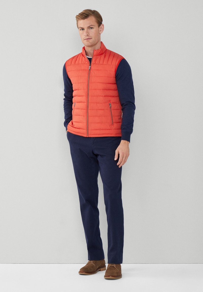Gilet rembourré rouge avec des coutures verticales, chemise à manches longues bleu marine en dessous, pantalons foncés et chaussures en suède marron. Poches avec fermeture éclair visibles.