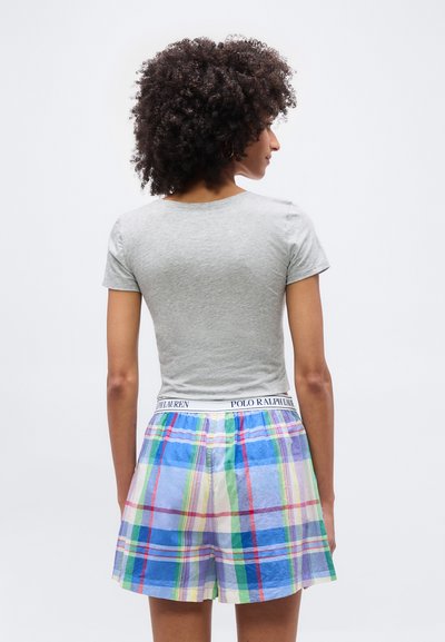Γυναίκα με σγουρά μαλλιά που φοράει ανοιχτό γκρι κοντομάνικη μπλούζα και πολύχρωμο καρό σορτσάκι με ζώνη Polo Ralph Lauren, γυρισμένη προς τα πίσω.