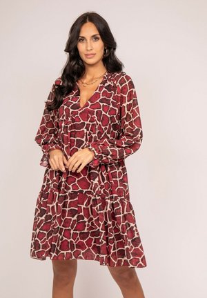 Femme aux longs cheveux foncés portant une robe ample à manches longues rouge et beige avec un imprimé girafe, debout devant un fond uni.