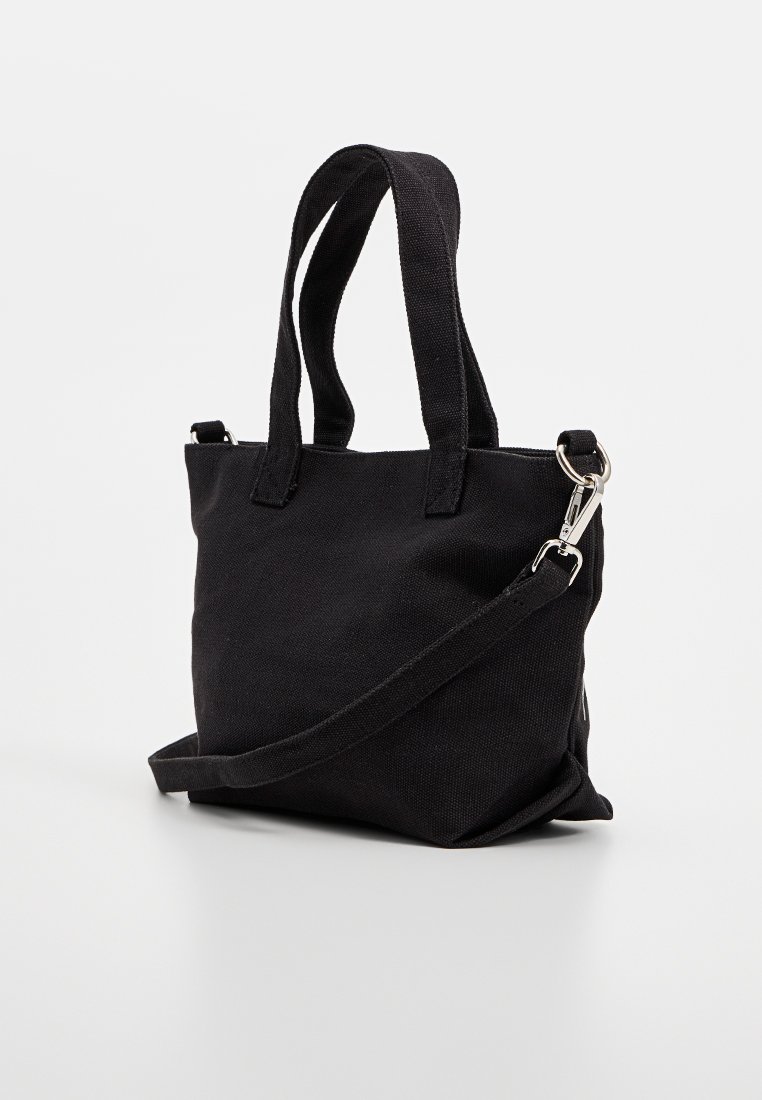 Sac fourre-tout noir en toile avec deux courtes poignées, une sangle d'épaule amovible et un détail en métal argenté. Design simple et structuré.
