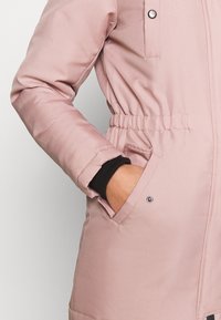 Rosa vadderad jacka med en slät textur, avsmalnande midja och sidofickor. Har tryckknappar och elastiska manschetter.