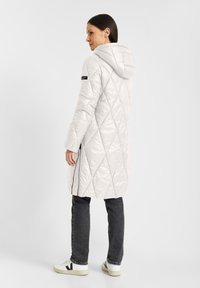 Lichte, gewatteerde, lange winterjas in lichtgrijs met een capuchon, voorzien van een diagonaal patroon en een zijrits. Gepaarde met donkere jeans en sneakers.