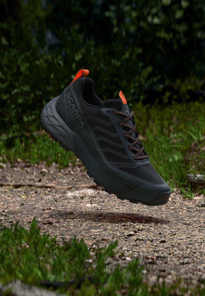 Μαύρο και γκρι παπούτσι τρεξίματος Scarpa με πορτοκαλί λεπτομέρειες, αιωρούμενο στον αέρα πάνω από μονοπάτι με χαλίκι και γρασίδι σε εξωτερικό περιβάλλον.