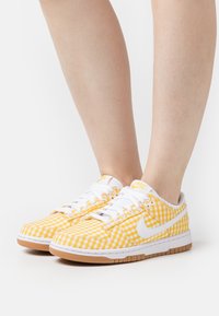 Sneakers a quadretti gialli e bianchi con tomaia in tessuto strutturato, lacci bianchi e suola in gomma color biscotto. Logo Nike visibile sui lati.