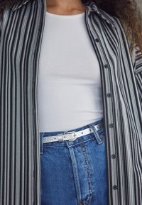Chemise à rayures noires et blanches avec boutons sur un débardeur blanc côtelé, associée à un jean en denim bleu taille haute et une ceinture argent métallique.